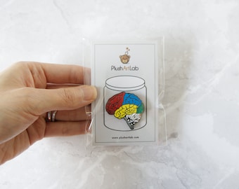 Könnte beinhalten: Eine farbige Gehirnstifte aus Emaille in einer durchsichtigen Plastiktüte. Die Stifte ist rot, gelb, blau und grün. Die Stifte befindet sich auf einer weißen Karte mit dem Text "PlushArtLab" und "www.plushartlab.com".