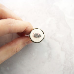 May include: A small, round ring with a white fabric center featuring an embroidered gray mouse with pink ears. The ring is made of metal with a dark brown finish.