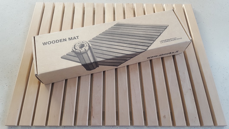 May include: A brown cardboard box with the words "WOODEN MAT" printed on the front. The box contains a wooden mat that is rolled up and tied with a rope. The mat is made of light brown wood and has a natural finish. The box also has the website address "www.almeks.lt" printed on the front.