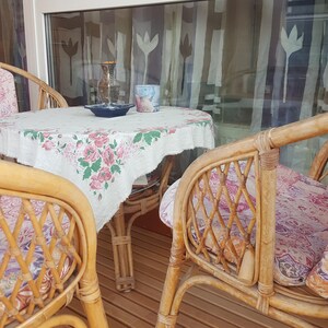 May include: Two wicker chairs with floral patterned cushions sit around a small table with a floral tablecloth. The table has a vase, a cup, and a plate on it. The chairs and table are on a wooden deck.