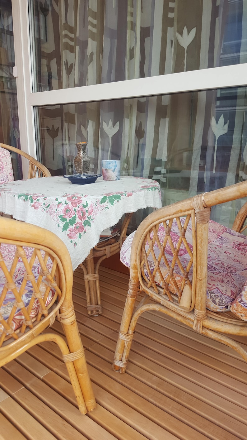 May include: Two wicker chairs with floral patterned cushions sit around a small table with a floral tablecloth. The table has a vase, a cup, and a plate on it. The chairs and table are on a wooden deck.
