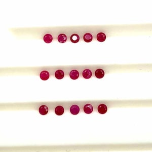 May include: A collection of small, round, faceted ruby gemstones. The rubies are a deep red color and are arranged in three horizontal rows against a white background. The gemstones are of varying sizes and clarity.