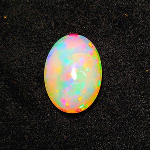 May include: Oval-shaped opal gemstone with a vibrant display of colors, including pink, green, yellow, and blue. The gemstone has a milky white base and is set against a dark, textured background.