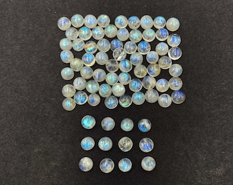 Op de afbeelding: Een verzameling ronde, gepolijste maansteen cabochons. De stenen zijn doorschijnend met een melkachtige witte basis en vertonen een blauwe adularescentie. De edelstenen zijn gerangschikt op een donkergrijze ondergrond.