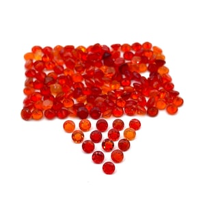 May include: A collection of faceted, round gemstones in shades of red and orange. The gems are scattered across a white surface, with a concentration of stones forming a rectangular shape and a downward-pointing triangle.