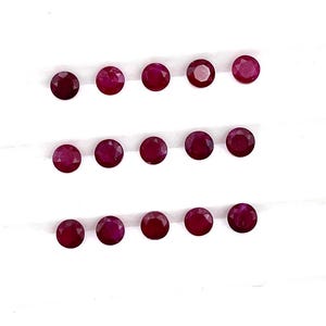 Puede incluir: Una colección de 15 piedras preciosas de rubí redondas y facetadas dispuestas en tres filas sobre una superficie blanca.