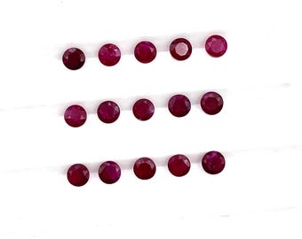 May include: A collection of 15 round, faceted ruby gemstones arranged in three rows on a white surface.