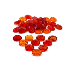 May include: A collection of faceted, oval-shaped gemstones in shades of red and orange. The gems are scattered on a white surface, with varying degrees of transparency and brilliance. The gemstones are likely for jewelry making or crafting.
