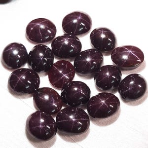May include: A collection of polished, oval-shaped, dark red gemstones. Each stone has a star-like pattern across its surface. The gems are arranged on a white surface, showcasing their deep color and reflective qualities. These are likely star garnet gemstones.