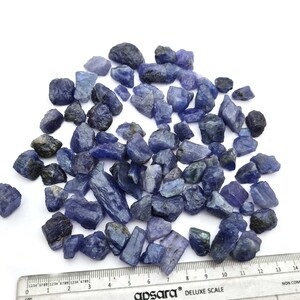 Op de afbeelding: Een hoop ruwe, blauw-paarse edelstenen, sommige met een lichtgroene tint, verspreid over een wit oppervlak. Een liniaal is zichtbaar aan de onderkant van de afbeelding, met markeringen in centimeters.