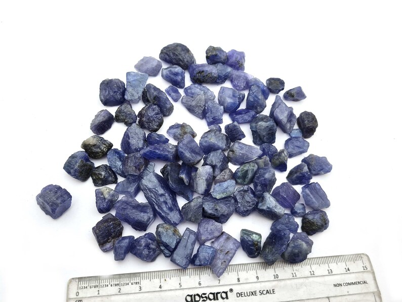 Op de afbeelding: Een hoop ruwe, blauw-paarse edelstenen, sommige met een lichtgroene tint, verspreid over een wit oppervlak. Een liniaal is zichtbaar aan de onderkant van de afbeelding, met markeringen in centimeters.
