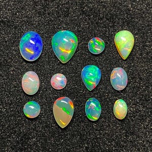 May include: Eleven opals of different shapes, including oval, teardrop, and round, are displayed. The opals show a variety of colours, such as blue, green, yellow, and red, with iridescent flashes. The gemstones are set against a black background.