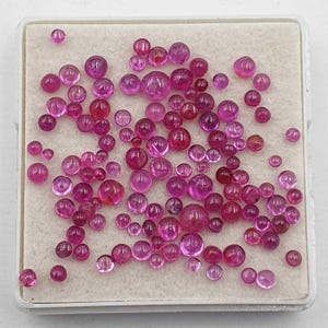 May include: A collection of numerous small, round, cabochon-cut gemstones in various shades of vibrant pink and deep red. These polished, translucent stones are arranged in a scattered pattern on a square, off-white, felt-lined display tray.
