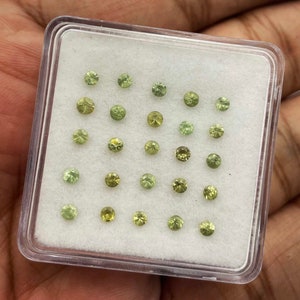 Puede incluir: Primer plano de una pequeña caja de plástico llena de 25 piedras preciosas verdes sueltas. Las piedras preciosas están dispuestas en filas y todas son aproximadamente del mismo tamaño.