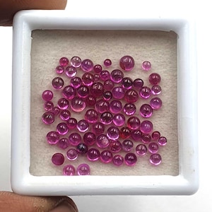 May include: A collection of small, round, cabochon-cut rubies in varying shades of deep pink and red. These loose gemstones are displayed inside a square white plastic storage box with a light-colored, textured foam insert holding them in place.