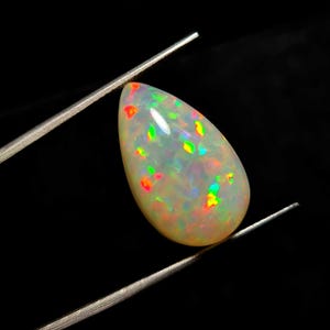 May include: A pear-shaped opal gemstone with a milky white base color. The opal displays vibrant flashes of color, including red, green, and yellow, held by metal tweezers against a black background. The gemstone is polished and smooth.