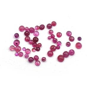 Op de afbeelding: Een verzameling kleine, ronde, losse kralen in diverse tinten roze, magenta en dieprood. Deze gepolijste, bolvormige kralen variëren in grootte en vertonen doorschijnende tot ondoorzichtige kwaliteiten, verspreid over een wit oppervlak.