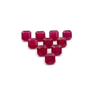 May include: A collection of ten loose, faceted, octagonal-cut rubies arranged in a triangular formation. Each gemstone displays a deep, saturated magenta-red colour with visible internal facets that reflect light across the surface.