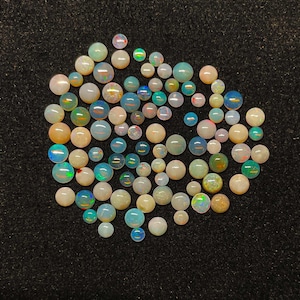 Puede incluir: Una colección de piedras preciosas de ópalo pulidas en varios tamaños y colores, incluyendo blanco, azul, verde y naranja. Los ópalos son redondos y se muestran sobre un fondo negro.
