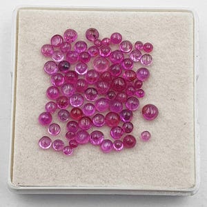 May include: A collection of numerous small, round, cabochon-cut rubies in varying shades of vibrant pink and deep red. These polished gemstones are arranged in a cluster on a square, off-white, felt-lined display tray with a white plastic frame.
