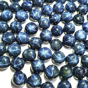 May include: A collection of blue star sapphires, each with a six-pointed star in the center. The sapphires are round and cabochon cut, and they are arranged in a random pattern.