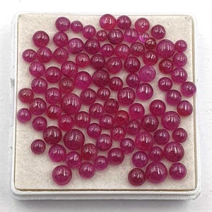 May include: A collection of numerous small, round, polished ruby gemstones in varying shades of deep pink and red. The stones are arranged in a cluster on a square, off-white display tray with a raised border, showcasing their smooth surfaces.