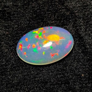 May include: An oval-shaped opal gemstone with a translucent, milky white base. The opal displays vibrant flashes of color, including red, green, and yellow, scattered throughout its surface. The gemstone is set against a dark, textured background.