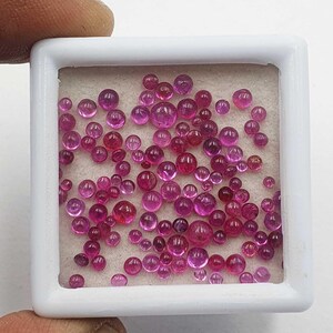May include: A collection of numerous small, round, cabochon-cut pink rubies displayed inside a square white plastic storage box. The gemstones vary in size and exhibit a translucent, vibrant magenta-pink hue with smooth, polished surfaces.