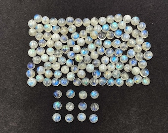 Op de afbeelding: Een verzameling ronde, doorschijnende maansteen cabochons met een melkachtige witte basis en flitsen van blauwe en iriserende kleur. De edelstenen zijn gerangschikt op een zwarte ondergrond.