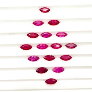 May include: A collection of fourteen loose, marquise-cut rubies arranged in a symmetrical, diamond-shaped pattern. Each gemstone features a vibrant, deep red hue with faceted surfaces that reflect light, showcasing a pointed, elongated shape.