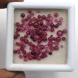 Peut inclure: Une collection de nombreuses petites pierres précieuses rondes et facettées de couleur rose et rouge foncé, disposées dans une boîte de présentation carrée blanche avec un insert en mousse claire. Les pierres varient en taille et en intensité.