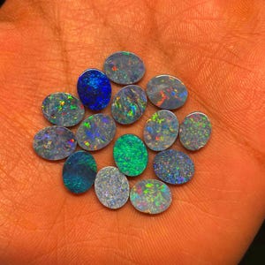 Puede incluir: Una colección de piedras preciosas de ópalo de forma ovalada en varios tonos de azul, verde y gris. Los ópalos tienen un efecto iridiscente brillante.