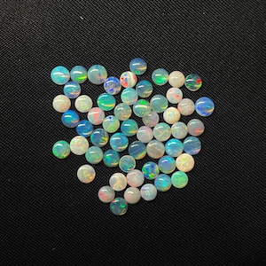 Puede incluir: Una colección de ópalos redondos y pulidos en varios colores, incluyendo blanco, azul, verde y rojo, están dispersos sobre una superficie negra. Los ópalos tienen un acabado brillante y muestran un juego de colores, creando una exhibición vibrante y llamativa.