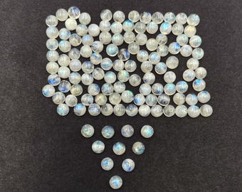 Op de afbeelding: Een verzameling ronde, doorschijnende maansteen cabochons met een subtiele blauw-witte iriserende glans. De edelstenen zijn verspreid over een donkere ondergrond, waarbij hun gepolijste, bolvormige vorm wordt getoond. Ideaal voor het maken van sieraden.