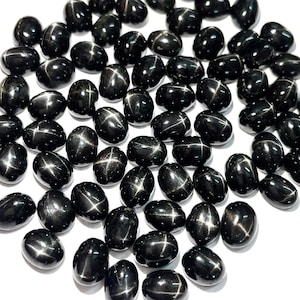 Pode incluir: Uma coleção de pedras preciosas de safira estrela preta polidas. Cada pedra em forma oval exibe um padrão proeminente em forma de estrela em sua superfície. As pedras preciosas estão dispostas em um fundo branco.