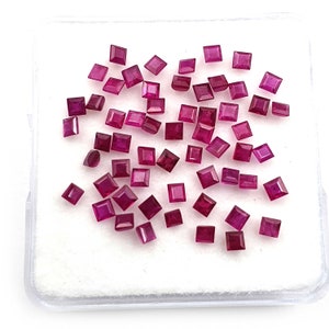 Puede incluir: Una colección de rubíes rojos oscuros de corte cuadrado. Las piedras preciosas están dispersas sobre una superficie blanca dentro de un recipiente de plástico transparente. Los rubíes varían ligeramente en tamaño y forma.