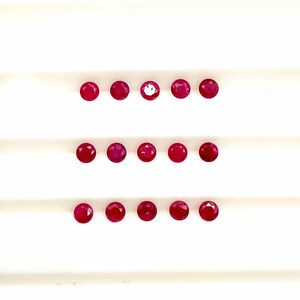 May include: Fifteen round, deep red gemstones are displayed in three horizontal rows against a white, subtly patterned background. The polished surfaces of the gemstones reflect light, highlighting their intense colour.