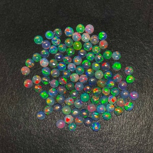 Può includere: Una collezione di pietre preziose di opale rotonde e iridescenti in varie tonalità di blu, verde e rosso. Gli opali sono sparsi su una superficie nera.