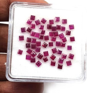 May include: A collection of small, square-cut, deep red gemstones, likely rubies, are displayed in a clear plastic container with a white interior. The gemstones are of varying shades of red, and are scattered across the white surface.