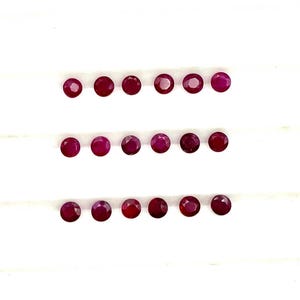 Puede incluir: Una fila de 18 pequeñas piedras preciosas redondas de color rojo dispuestas en tres filas de seis. Las piedras preciosas están dispuestas sobre un fondo blanco.