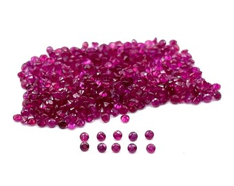 May include: A large pile of small, round, red ruby gemstones. The rubies are faceted and have a brilliant shine. There are also a few rubies arranged in a row below the pile.