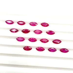 Puede incluir: Un conjunto de quince piedras preciosas sueltas, talladas en forma de marquesa, con un tono rojo rosado intenso y vibrante. Cada piedra presenta una superficie facetada que refleja la luz, dispuesta en cuatro filas horizontales sobre una superficie blanca estriada.