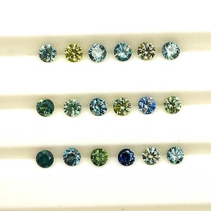 May include: A collection of small, round gemstones in a range of colours, including blues, greens, and yellows. The gems are arranged in neat rows, highlighting their facets and sparkle. The gemstones are set against a white background.