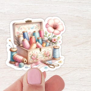 May include: A vintage wooden box filled with colourful spools of thread, a pair of scissors, and a pink flower. The box has a floral design and is surrounded by a white background.