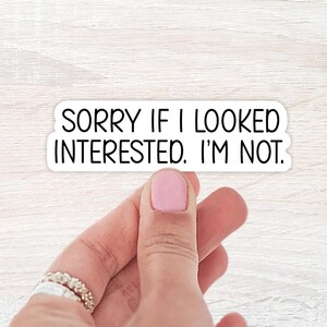 May include: A white sticker with the text "SORRY IF I LOOKED INTERESTED. I'M NOT." in black, held by a hand with a pink painted nail and a silver ring. The background is a light wood grain.