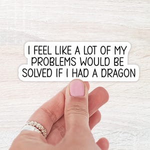 May include: A white sticker with black text that reads "I FEEL LIKE A LOT OF MY PROBLEMS WOULD BE SOLVED IF I HAD A DRAGON." The sticker is held by a hand with a ring.