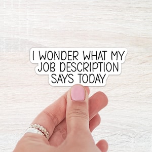 May include: A white sticker with black text that reads "I WONDER WHAT MY JOB DESCRIPTION SAYS TODAY". The sticker is held by a hand with a pink painted nail and a ring.