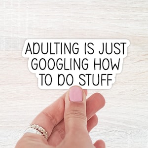 May include: A white sticker with black text that reads "ADULTING IS JUST GOOGLING HOW TO DO STUFF." The sticker is held by a hand with a pink painted nail and a silver ring. The background is a light wood grain.