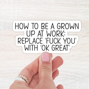 May include: A white sticker with black text that reads: "HOW TO BE A GROWN UP AT WORK: REPLACE 'FUCK YOU' WITH 'OK GREAT'". The sticker is being held by a hand with pink nail polish and rings.