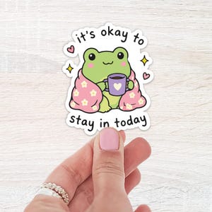 Peut inclure: Un autocollant découpé représentant une grenouille de dessin animé vert clair enveloppée dans une couverture rose à motifs de fleurs blanches, tenant une tasse violette avec un cœur blanc. Le texte indique, it's okay to stay in today.